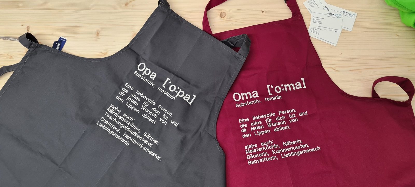 Eine graue und eine rote Schürze, bestickt mit Opa und Oma und einem langen Text